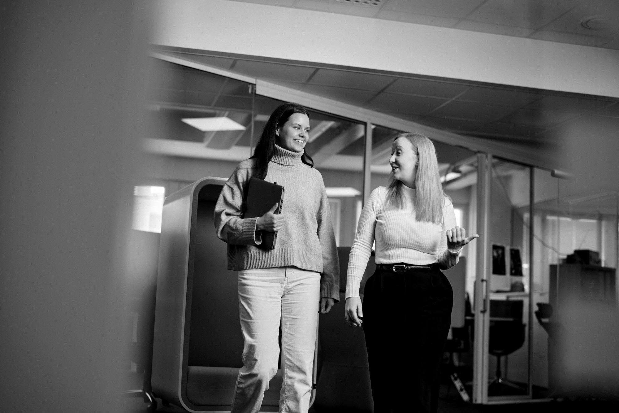 Kaksi naista kävelee toimiston aulassa. Toinen naisista kantaa tietokonetta.